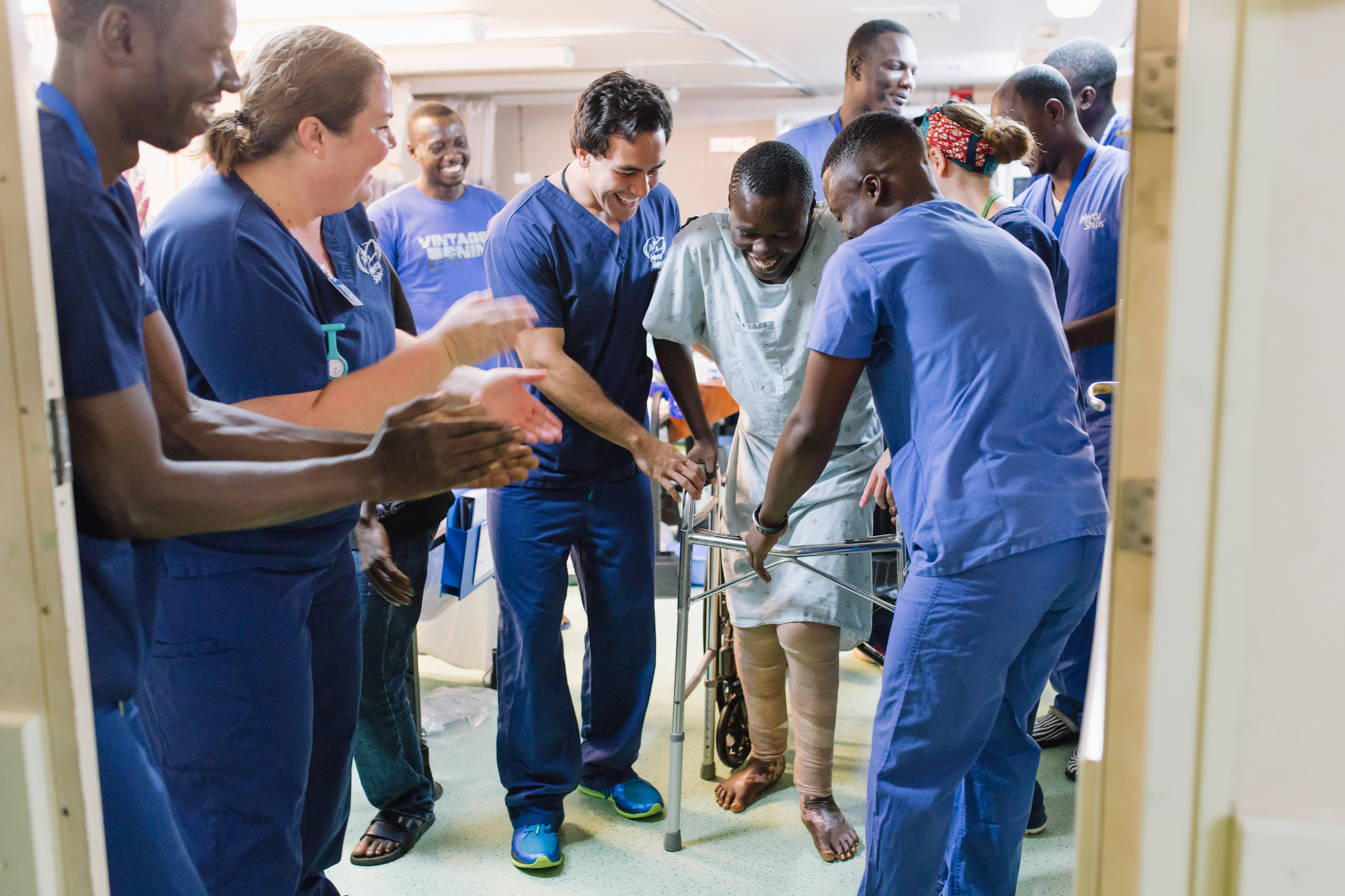 Ibrahima, plastics patient, walking for the first time in the ward after surgery with the help of Allan Kent, Physical Therapist, and others as his brother watches proudly in the background.